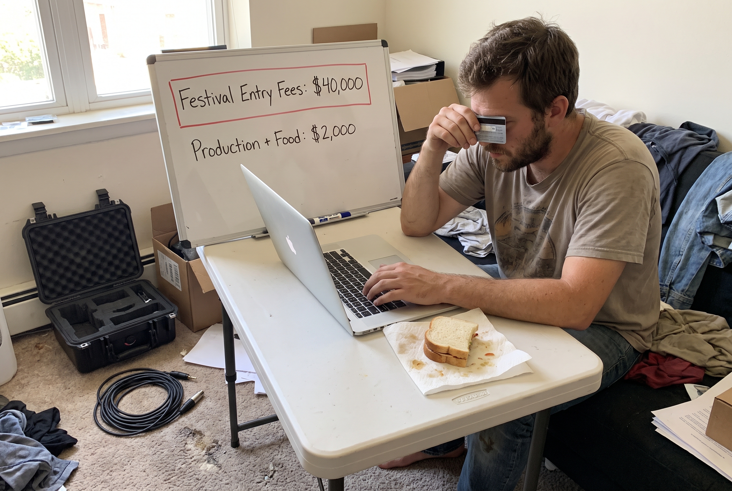 A man enters credit card details on a laptop next to a whiteboard outlining a $40,000 festival fee budget.