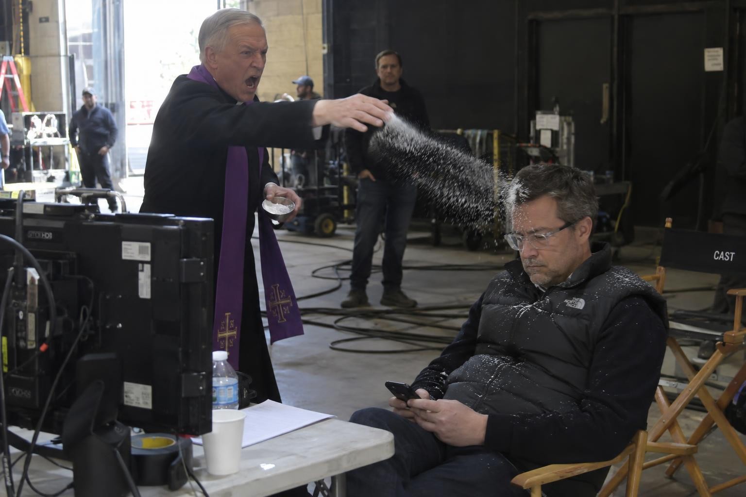 A Catholic priest aggressively throws white salt at a bored man in a quilted vest who is checking his phone next to film monitors.