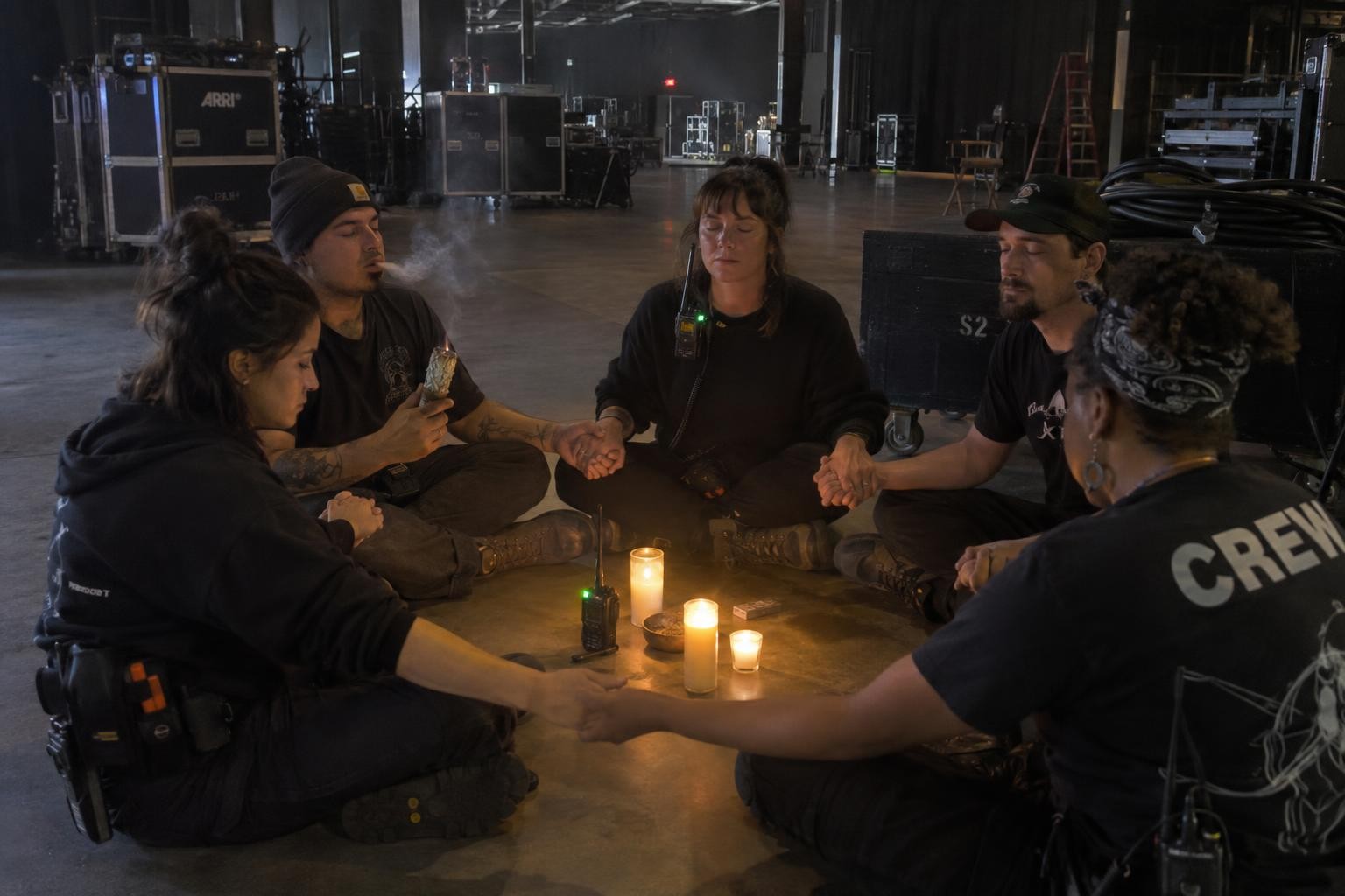 Five film crew members hold hands in a circle around lit candles, burning sage, and a walkie-talkie on a dark soundstage.