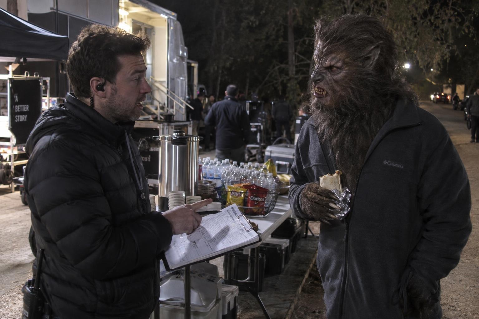 A film producer holding a clipboard talks to an actor in full werewolf makeup who is holding a burrito at a craft services table at night.