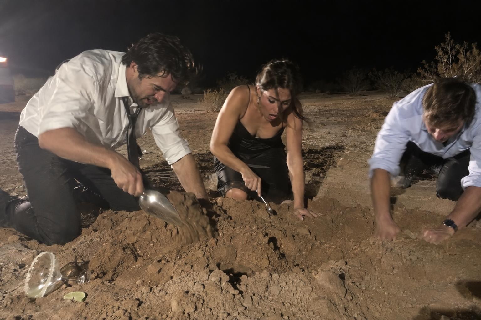 Three people in evening wear frantically dig into a mound of dirt at night using a tiny cocktail spoon and an ice scoop.