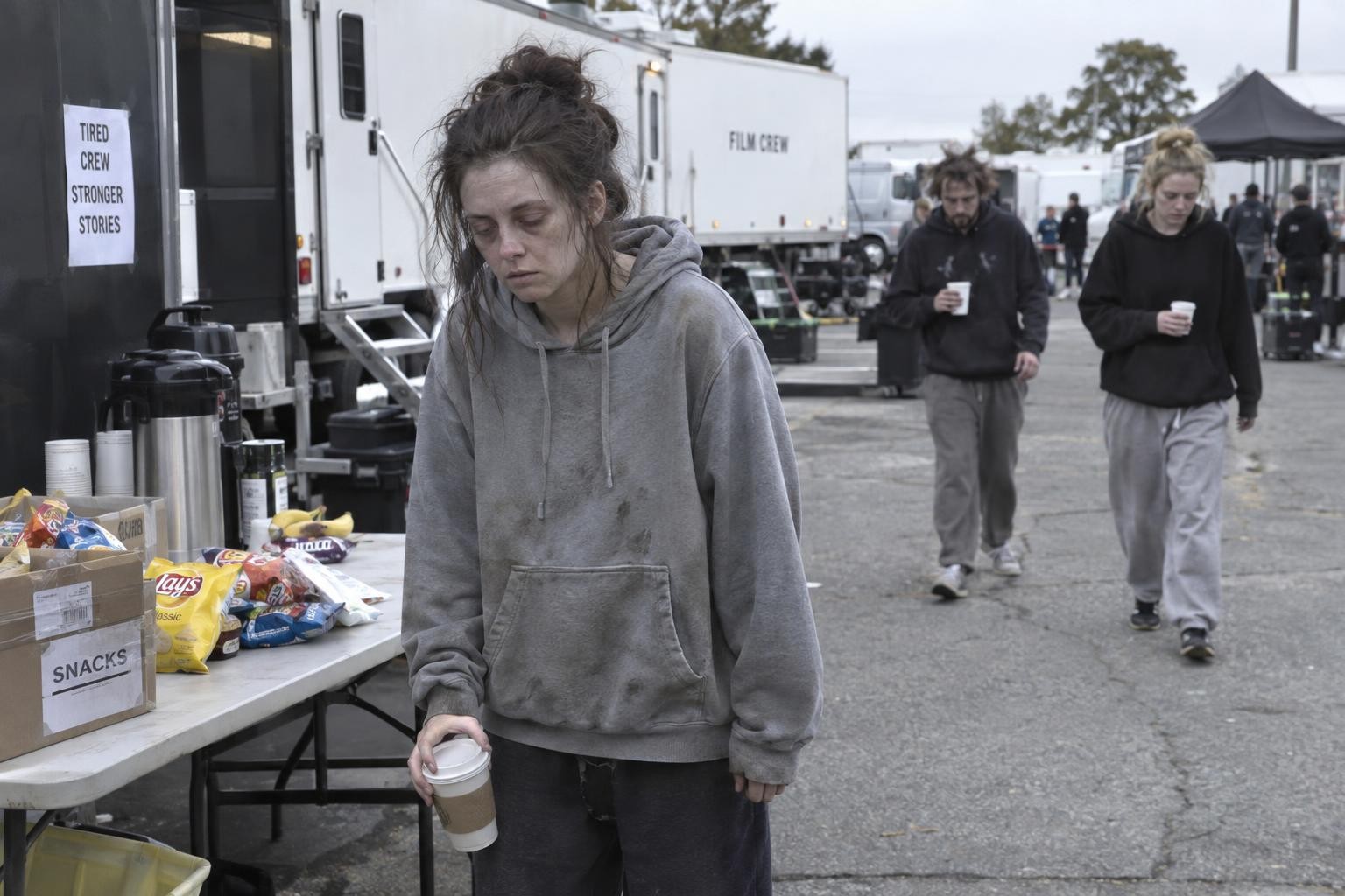 A pale, exhausted woman in a dirty hoodie stares blankly next to a snack table while two messy people shuffle behind her.