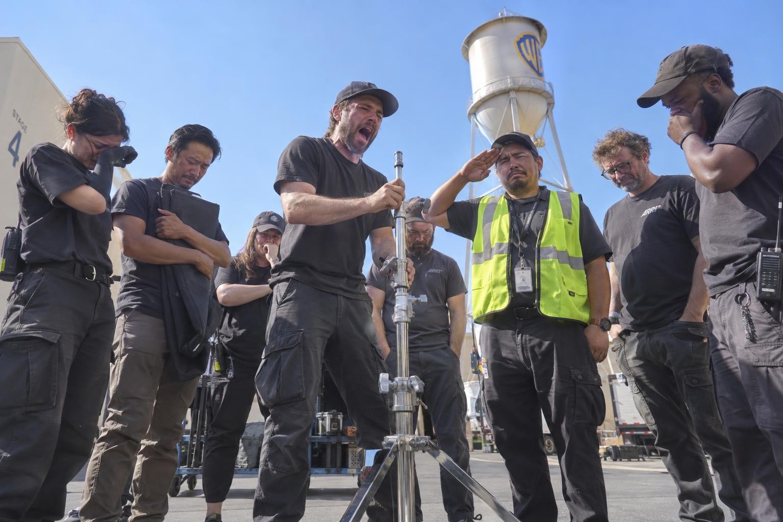 Devastated film crew members watch and salute as a man solemnly lowers a C-stand.